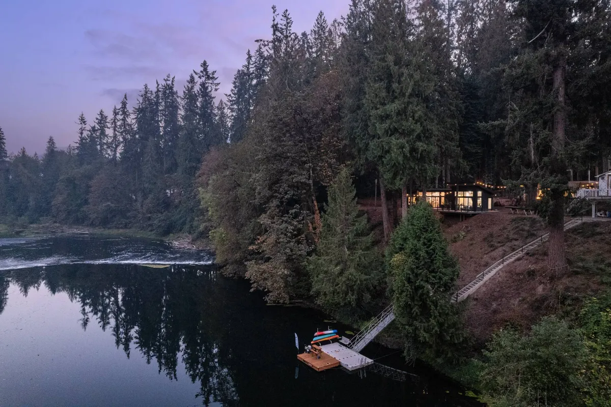 The image shows a serene riverside scene with the dock and colorful kayaks in the foreground, the house warmly illuminated in the background, and lush trees enclosing the area. The inviting mood suggests both daytime activity and relaxing evenings by the water.