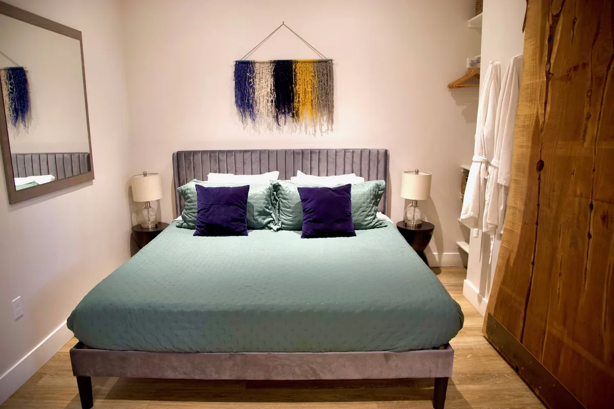 A calm and serene bedroom featuring a plush bed with neutral linens, tasteful wall art above the headboard, a large mirror, and matching nightstands. The natural light and uncluttered decor contribute to a sense of peace and relaxation.