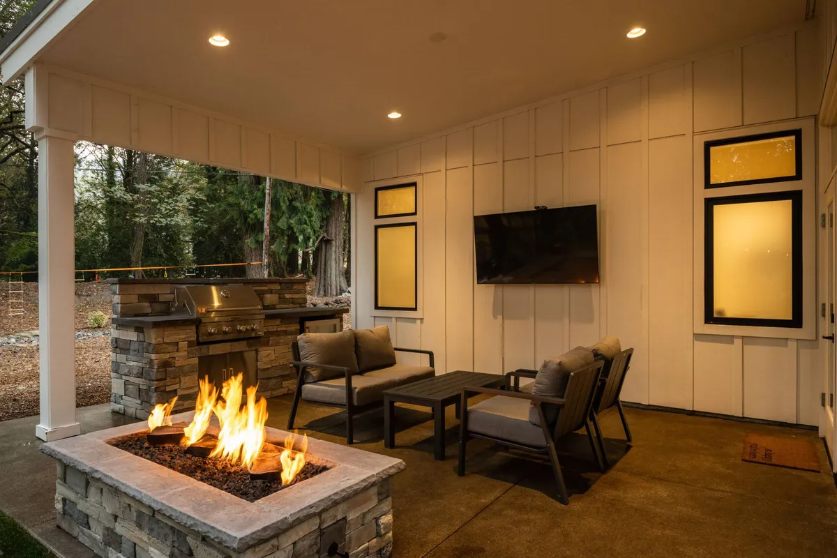 A welcoming outdoor lounge setup beneath a protective roof, featuring a prominent fire pit, comfortable seating, a built-in grill, and a large TV screen. The setup is ideal for entertaining, with a sense of warmth and togetherness, surrounded by natural greenery.