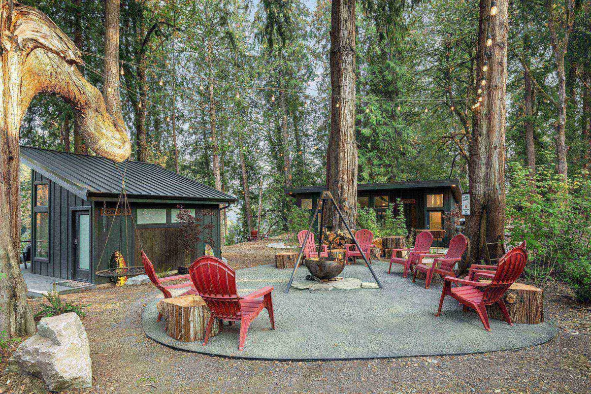 A central courtyard scene with a striking hanging iron fire pit as the focal point, surrounded by red chairs and whimsical tree sculptures amidst lush greenery. The area invites groups to gather, relax, and enjoy nature. The cozy arrangement hints at evenings spent by the fire, laughter, and friendly conversations.