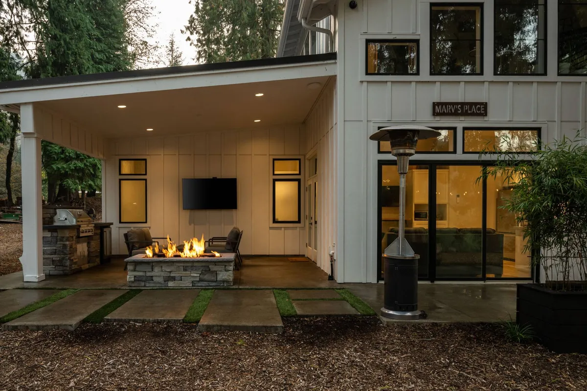 A spacious covered patio featuring a cozy fire-table at the center, surrounded by plush outdoor seating. The area includes a built-in grill for easy outdoor cooking, a mounted TV for entertainment, and overhead heaters, all set against a backdrop of towering forest trees. The sunset casts a warm glow, enhancing the relaxed, inviting atmosphere.