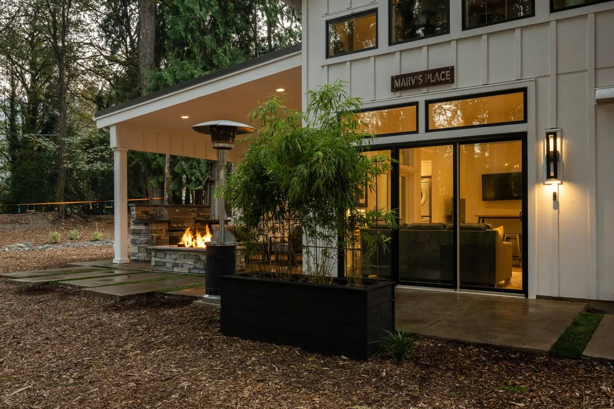 This exterior shot captures the inviting outdoor patio area at Marv’s Place, featuring a fire pit surrounded by comfortable seating, bamboo plants, an outdoor heater, and the prominent sign 'MARY'S PLACE' on the building. The large windows provide a visual link between the indoor and outdoor environments.