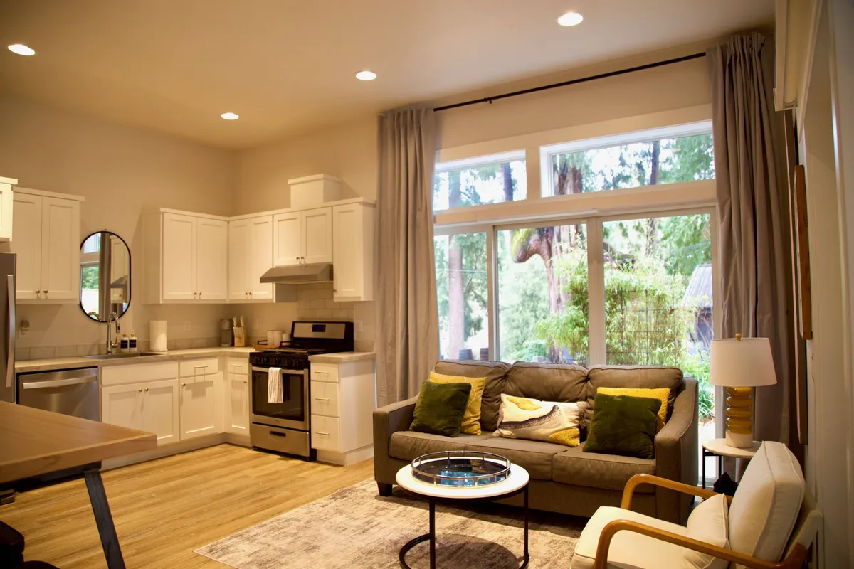 This image showcases the main living area with a plush sofa accented by decorative pillows, large windows that frame forest views, and a modern kitchen with visible appliances. Sunlight pours through the windows, creating a welcoming and tranquil atmosphere that blends indoor comfort with natural surroundings.
