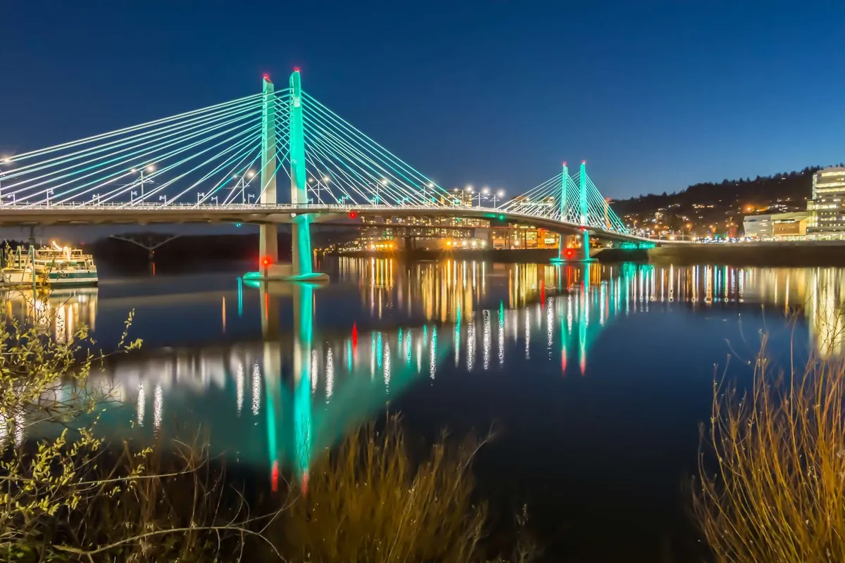 The image features a suspension bridge with city lights reflecting in the water, combining urban vibrancy with tranquil riverside scenery. The lively ambiance suggests nearby restaurants and nighttime activity, capturing the dynamic spirit of local exploration.