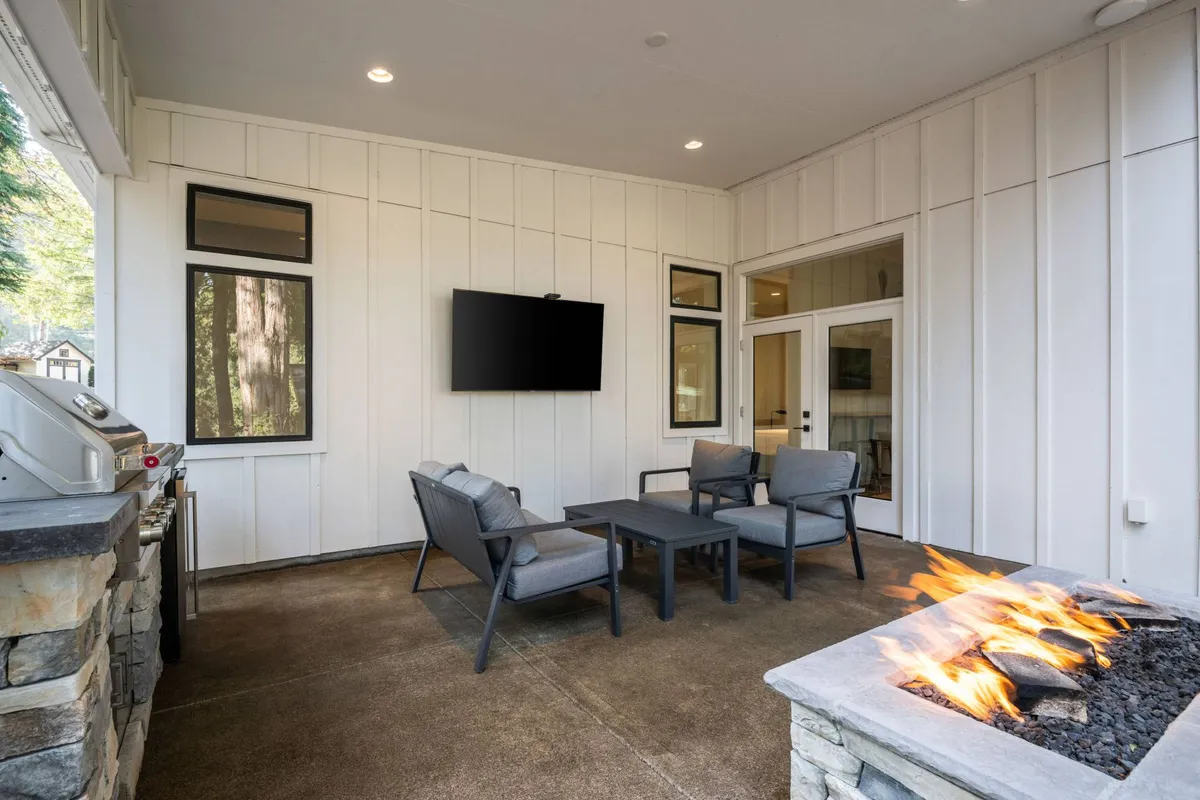 The image features a cozy outdoor living space with comfortable seating arranged around a glowing fire pit, all set beneath the open sky. An outdoor television invites guests to enjoy movies or sports under the stars, creating an inviting setting for relaxation and intimate gatherings. The backdrop showcases lush greenery, hinting at the property's tranquil, secluded location above the Tualatin River.