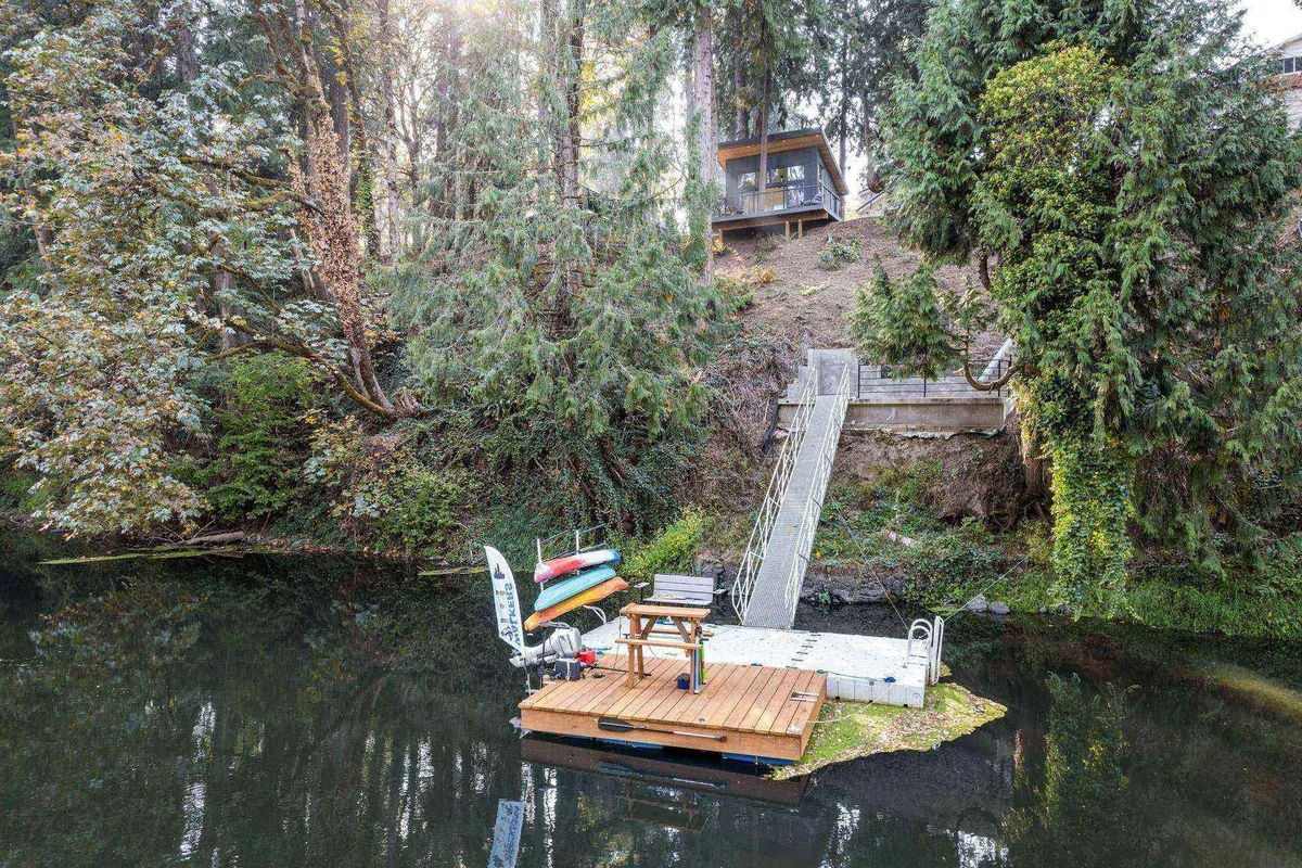 Here, a wooden dock extends over the calm river, with canoes and kayaks ready for adventure. Steps lead up to the house, tying the riverside experience directly to the living space. The image is suffused with tranquil, natural beauty, perfect for river access and enjoying peaceful moments on the water.
