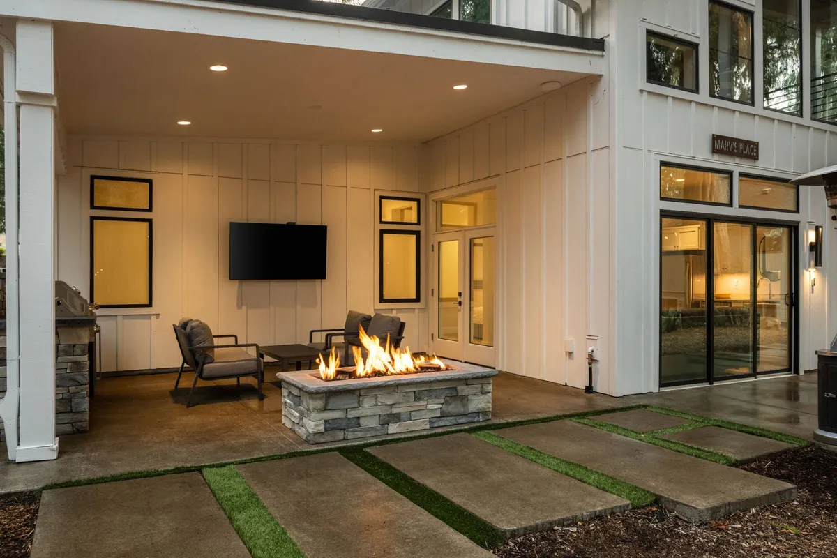 An inviting seating area on the patio with a fire pit, outdoor television, and comfortable chairs, all accessible via sliding glass doors that hint at the smooth transition between indoor and outdoor spaces. The setting is perfect for evening relaxation, movie nights, or gathering with friends and pets.