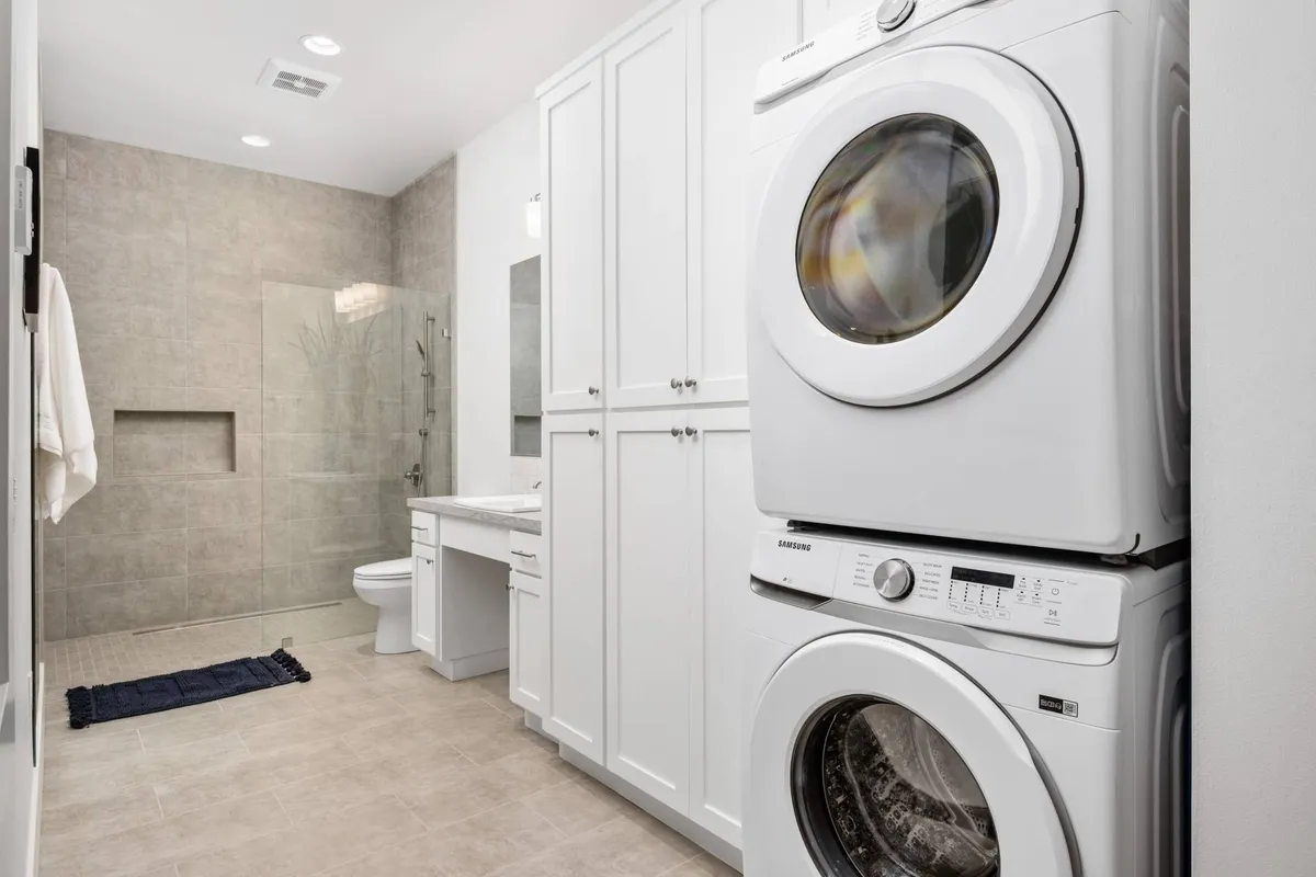 A modern and efficient bathroom/laundry area with a stacked washer and dryer, a walk-in shower with glass doors, and a sleek vanity with a sink. The space is tidy and thoughtfully laid out for multitasking and comfort.
