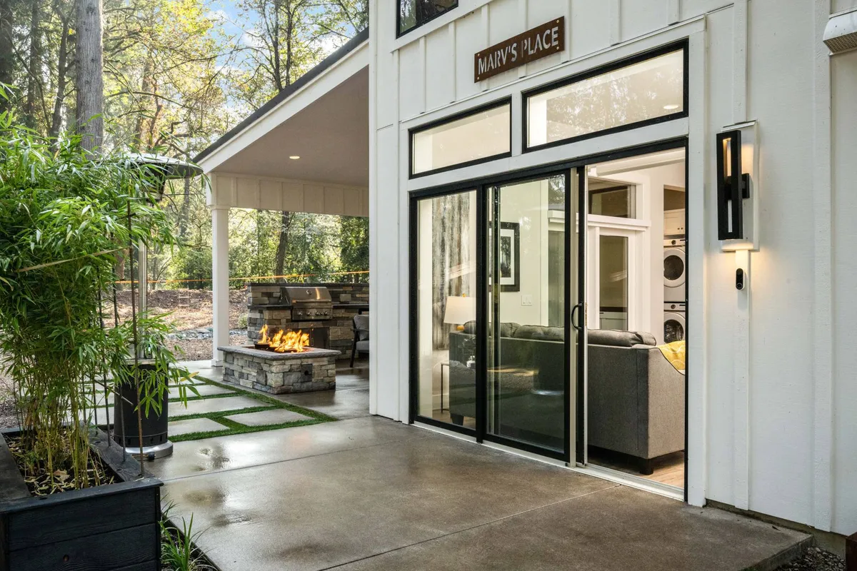 A welcoming exterior setting featuring an outdoor fireplace, glass sliding doors leading into the house, a landscaped pathway, and lush bamboo plants. The area appears inviting and is ideal for outdoor gatherings or relaxing in nature.