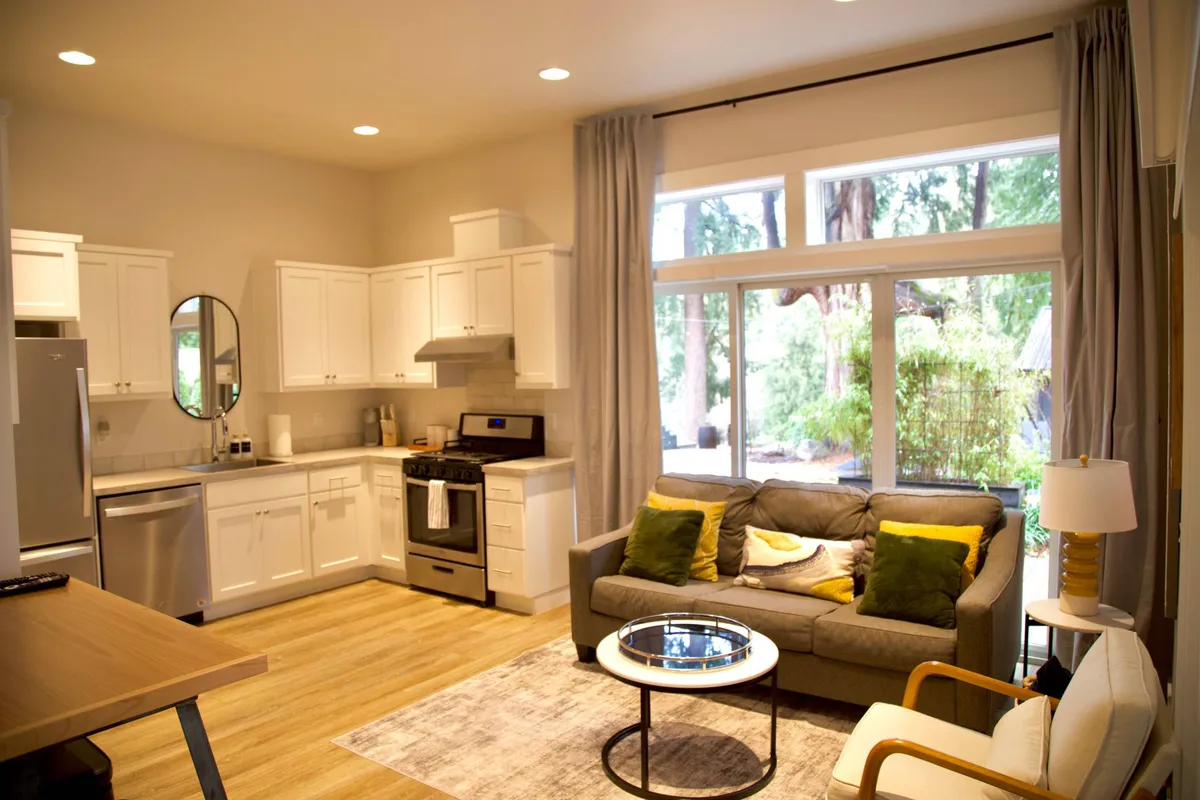 An inviting shot featuring the open-concept living, dining, and kitchen area. A cozy sofa faces large windows, with a dining table and modern kitchen appliances set in the background. The space is bathed in natural light, suggesting a warm, interconnected atmosphere ideal for both relaxation and socializing.